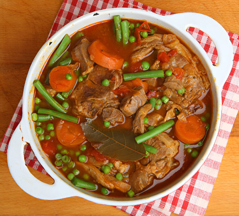 A hearty meat stew with carrots, green beans, peas, and bay leaves in a white serving dish.