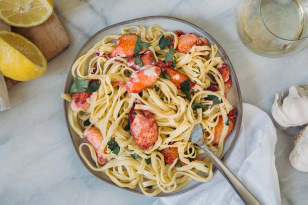 A plate of lobster fettuccine garnished with parsley on a marble surface, accompanied by lemon halves, garlic, and a glass of white wine.
