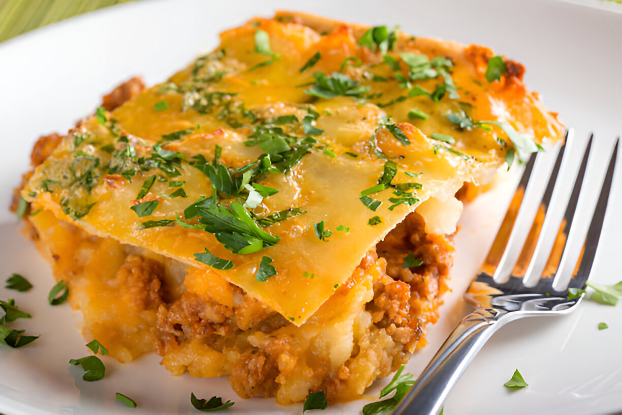 A close-up of a slice of lasagna topped with melted cheese and garnished with fresh parsley on a white plate alongside a fork.