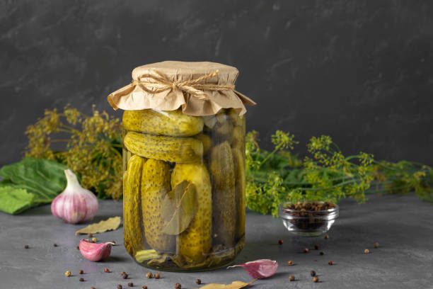 A jar of pickled cucumbers with a paper cover, surrounded by garlic, herbs, and spices on a gray surface.