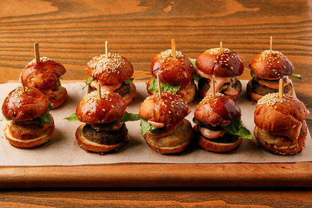 A selection of mini burgers on a wooden platter, topped with sesame seeds and garnished with leafy greens