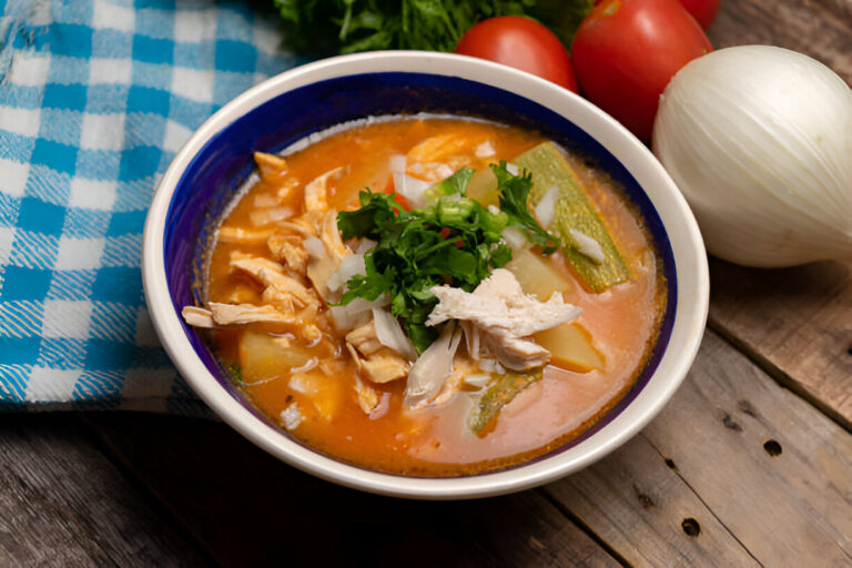 A bowl of colorful, hearty soup with chicken, vegetables, and herbs, resting on a wooden surface with tomatoes and an onion in the background.