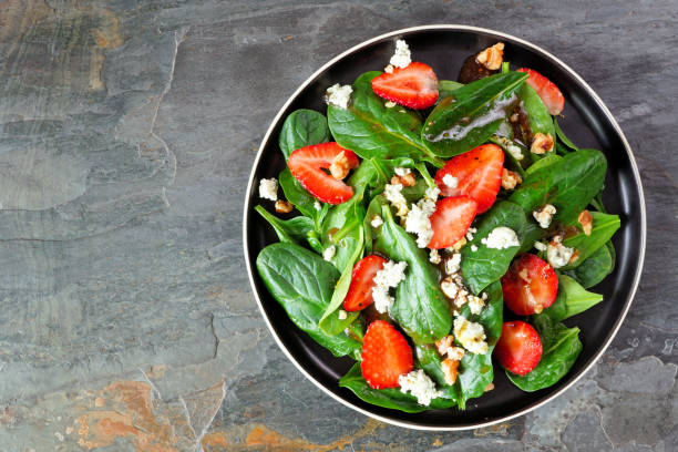 A fresh salad featuring spinach, strawberries, and goat cheese served in a black bowl on a textured stone surface.