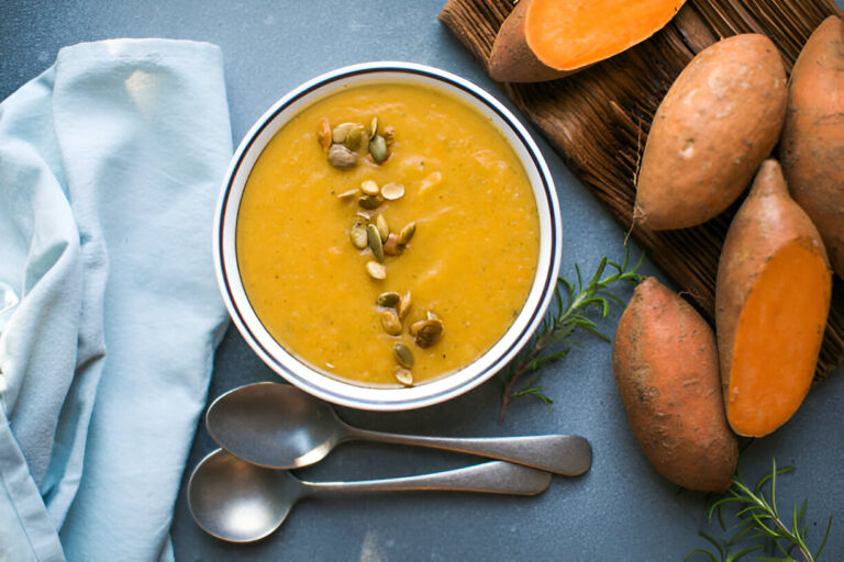 A bowl of rich yellow sweet potato soup garnished with pumpkin seeds, surrounded by whole sweet potatoes and silver spoons.