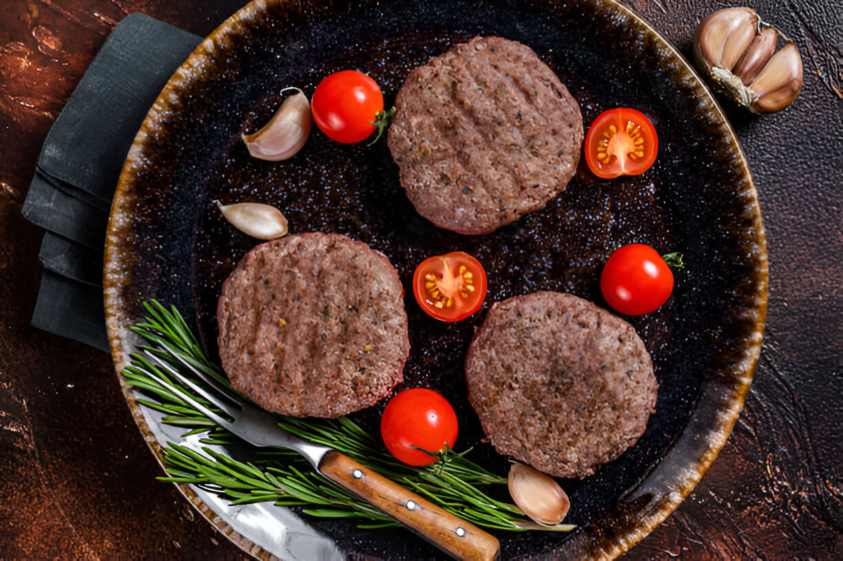 Three beef patties arranged on a dark plate with cherry tomatoes and garlic cloves.
