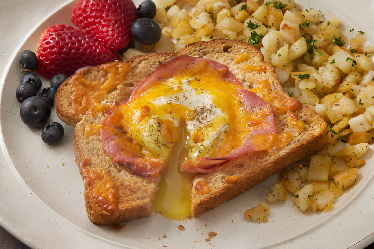 A plate of breakfast featuring toasted bread with melted cheese, ham, and a runny egg in the center, accompanied by diced potatoes and fresh berries.