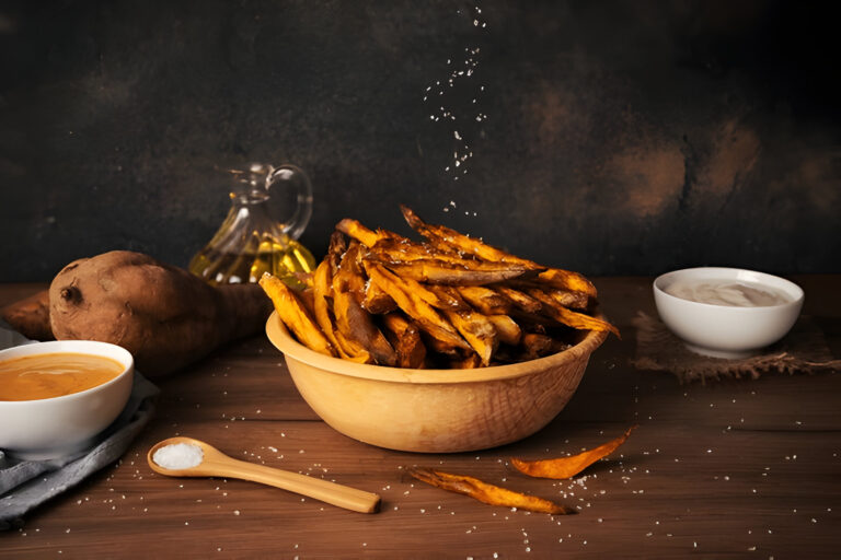 A wooden bowl filled with crispy sweet potato fries, garnished with salt, alongside a sweet potato, dipping sauces, and olive oil.