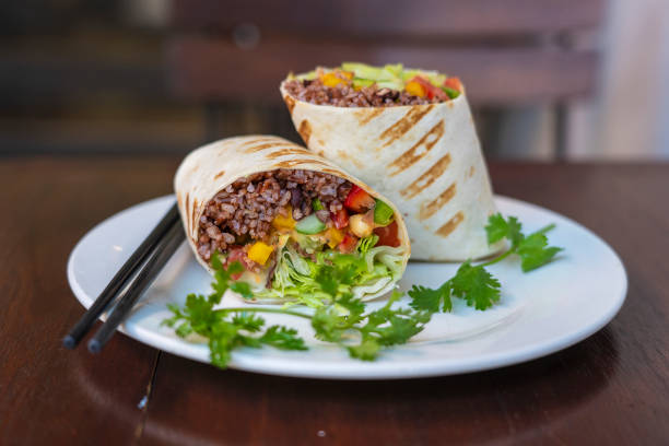 Two colorful burritos filled with rice, vegetables, and wrapped in a grilled tortilla, served on a white plate with fresh cilantro and chopsticks.