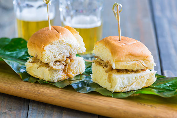 Two pulled pork sliders on a wooden platter with green leaves and beer in the background.