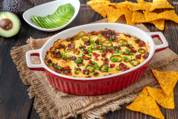 A delicious baked dish in a red casserole dish topped with bacon, jalapeños, and green onions, served with tortilla chips and slices of avocado.