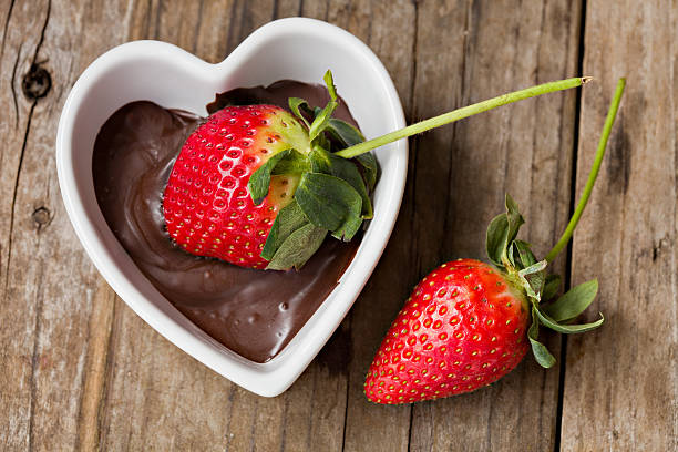 A fresh strawberry partially dipped in chocolate sauce, resting in a heart-shaped dish, with another strawberry beside it on a wooden table.