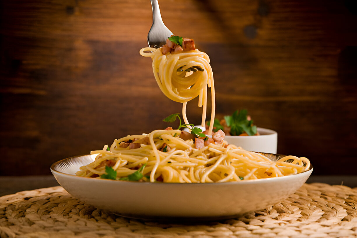 A fork lifting spaghetti pasta from a plate with bacon and herbs, set against a wooden background.