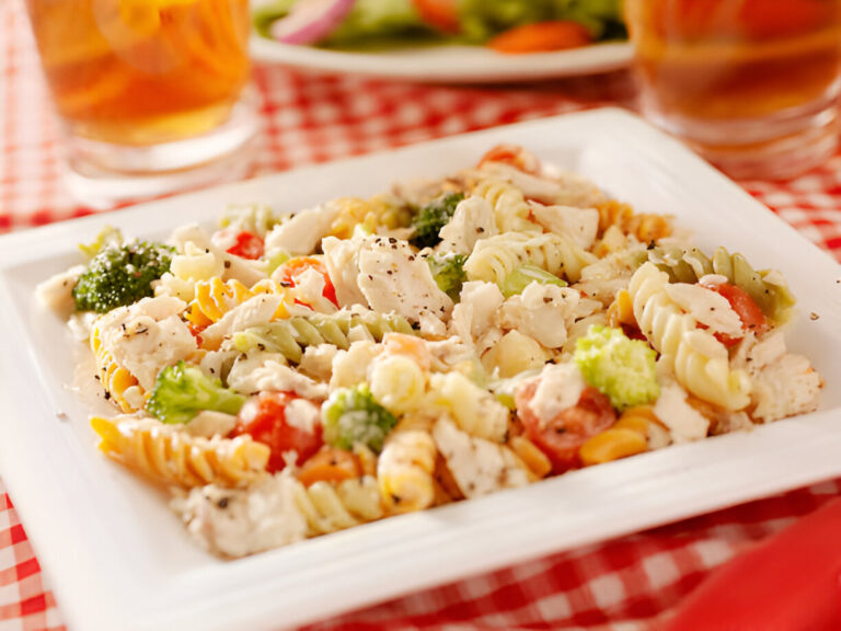 A plate of colorful pasta salad with broccoli, tomatoes, and chicken on a checkered tablecloth.