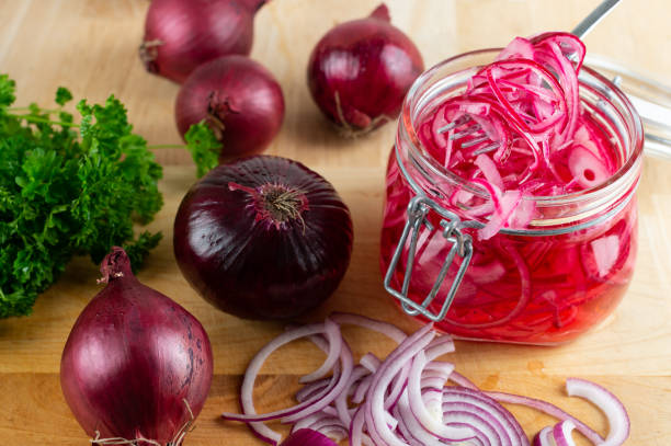 A jar of pickled red onions surrounded by fresh red onions and parsley on a wooden surface.