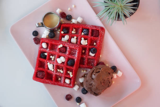 Two red waffles topped with blueberries, raspberries, mini marshmallows, and served with chocolate ice cream and syrup.