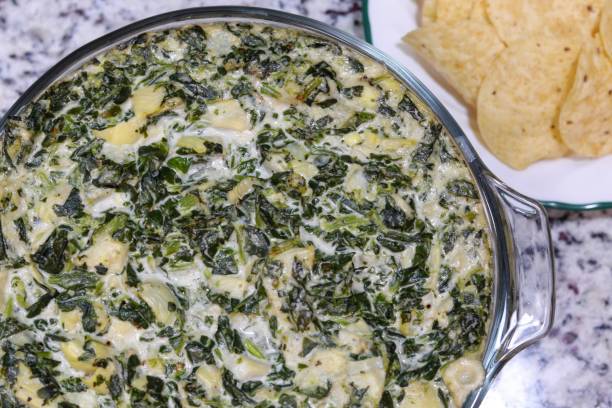 A creamy spinach dip served in a glass dish next to a plate of tortilla chips.