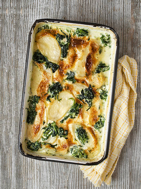 A baked dish of scalloped potatoes with spinach, featuring golden-brown top layers and creamy sauce, placed on a rustic wooden surface beside a checkered cloth.