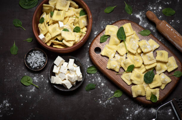 A wooden board and a bowl filled with fresh ravioli, sprinkled with flour and garnished with spinach leaves. A small bowl of feta cheese and another bowl of sea salt are placed nearby.