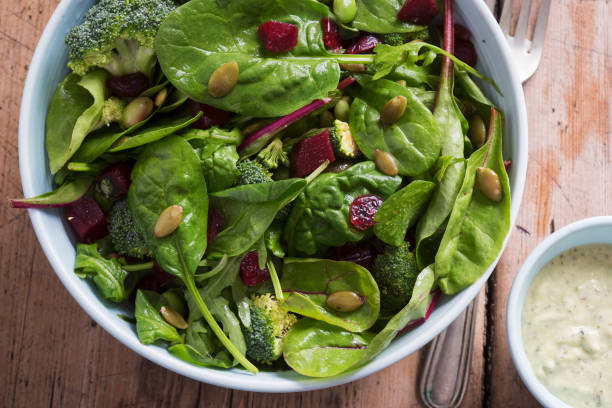 A fresh green salad with spinach, broccoli, and beets in a white bowl next to a small dish of dressing.