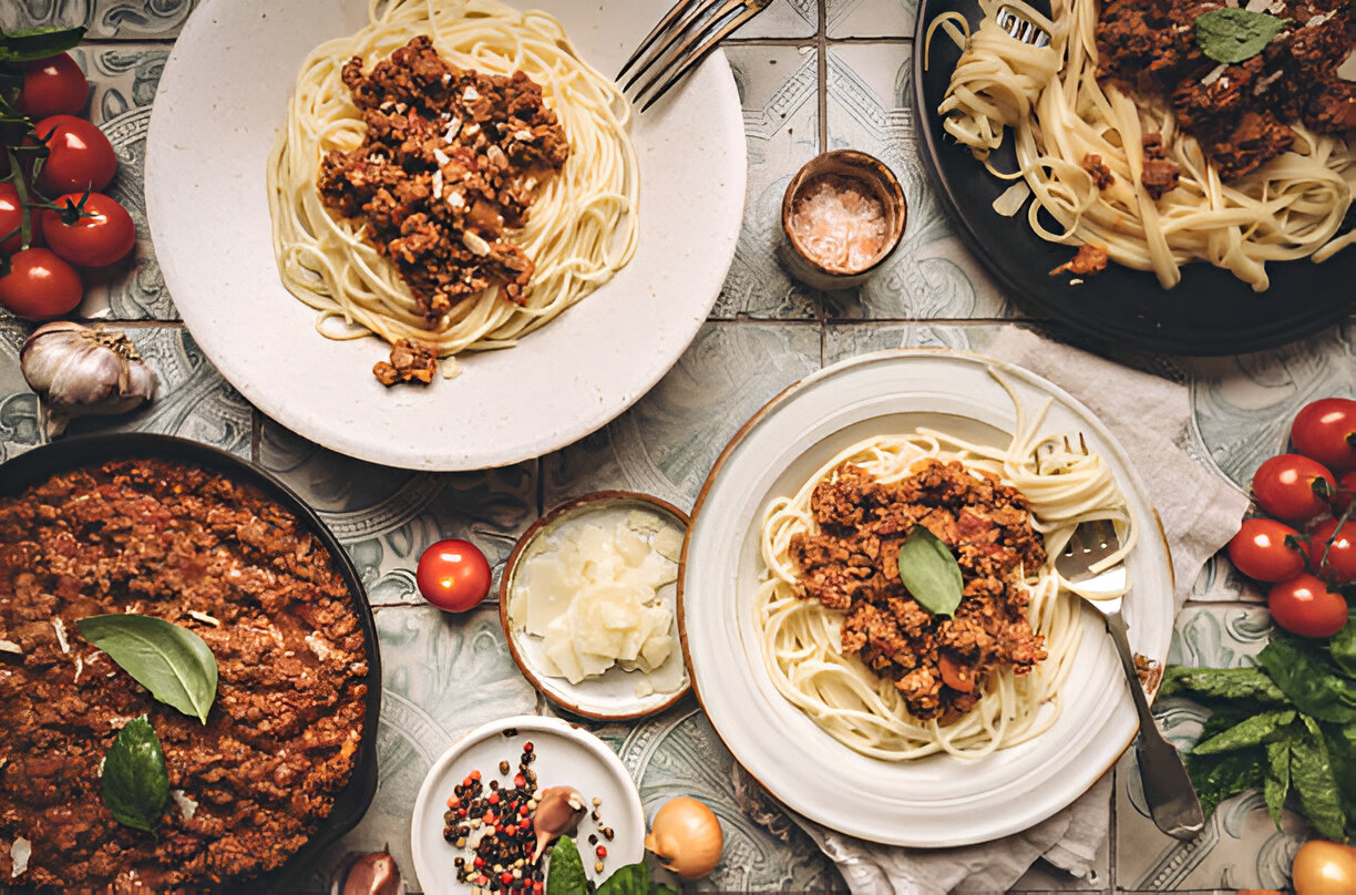 A beautifully arranged plate of spaghetti topped with meat sauce, accompanied by fresh tomatoes, garlic, and herbs on a decorative tiled surface.