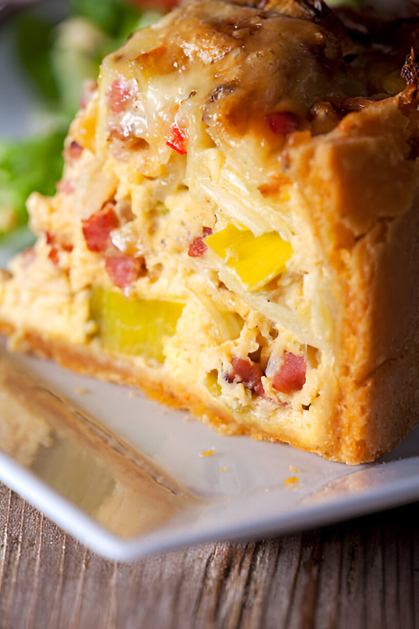 A slice of savory pie on a white plate, showcasing layers of filling with ingredients like yellow peppers and bacon.