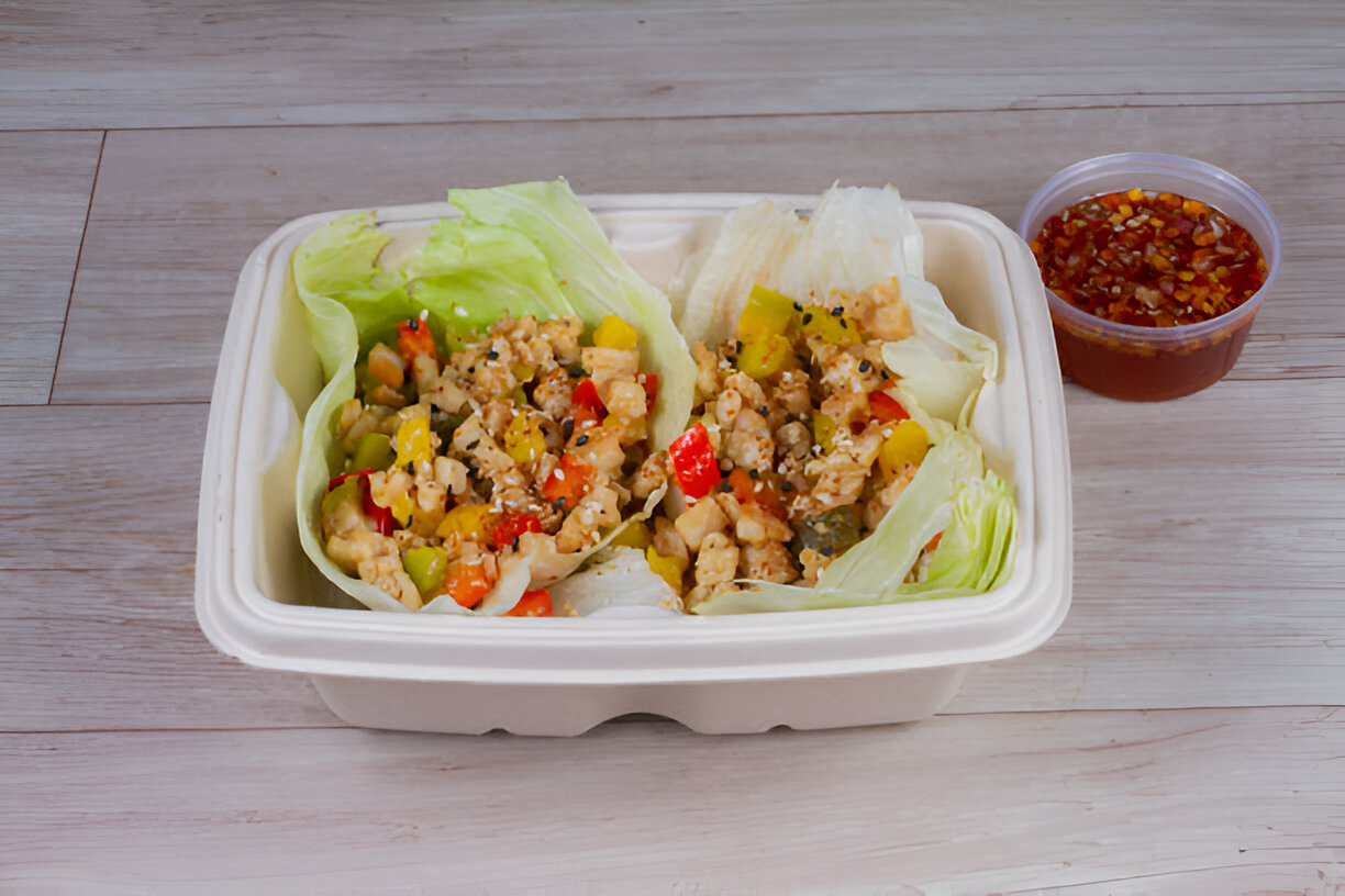 A container with lettuce wraps filled with a mixture of diced vegetables and protein, accompanied by a small container of sauce.