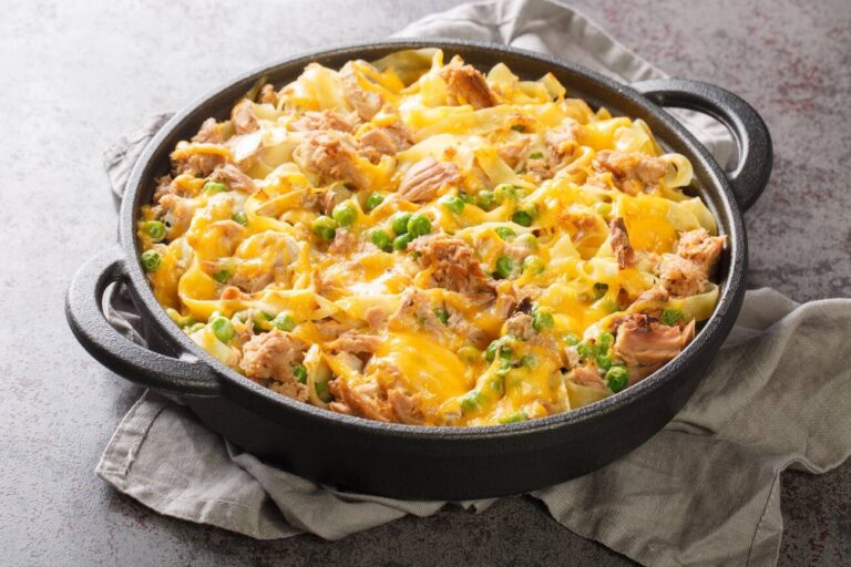 A close-up of a baked pasta dish with a golden cheese topping, mixed with tuna, peas, and egg noodles in a black serving dish.