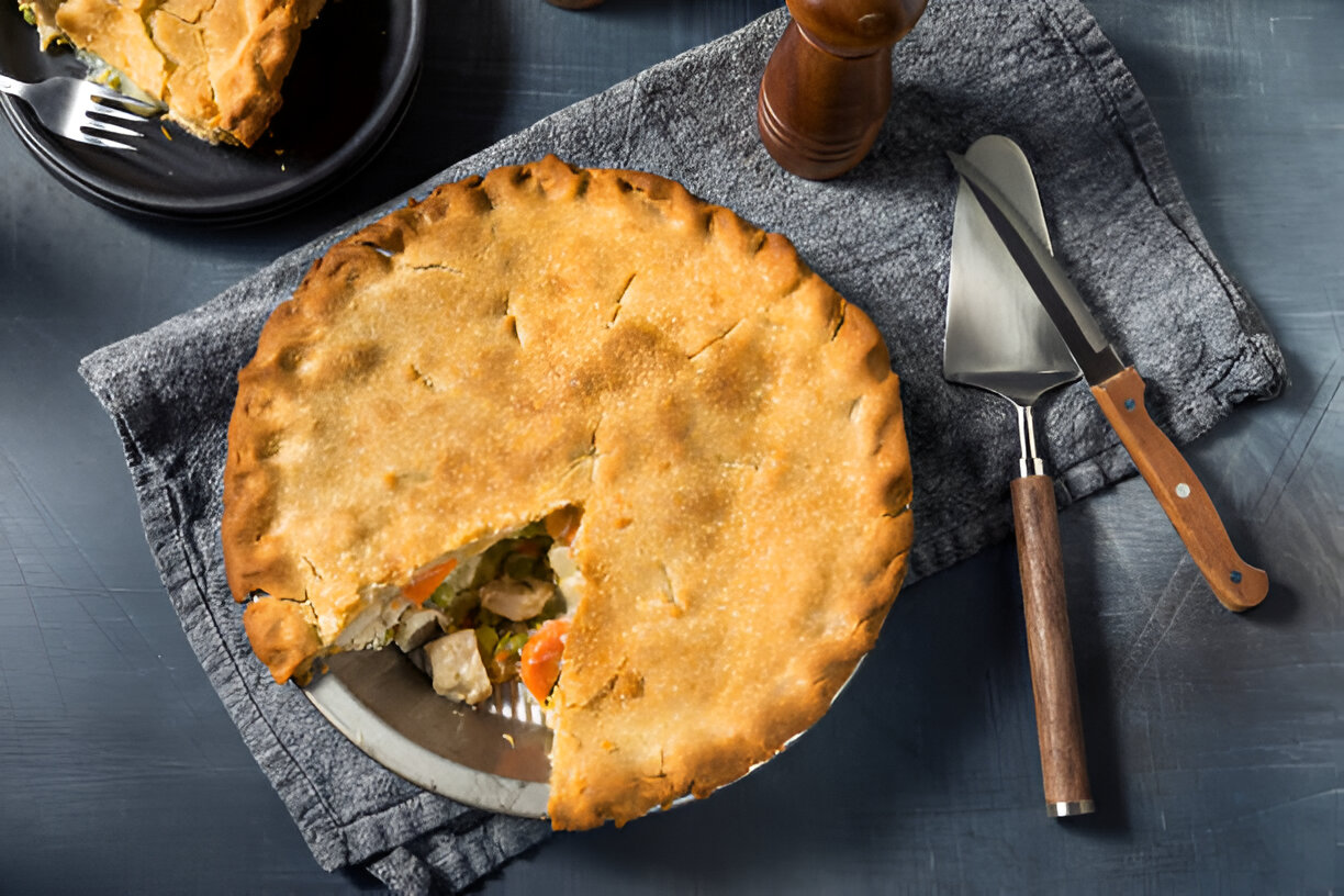 A freshly baked chicken pot pie with a golden crust, served with a slice on a black plate, set on a gray textured surface.