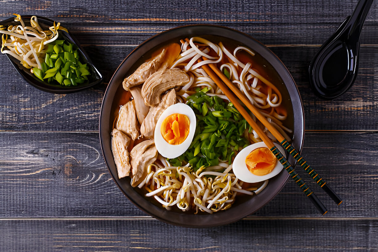 A bowl of delicious chicken noodle soup with sliced chicken, halved soft-boiled eggs, green onions, and bean sprouts served with chopsticks and a side of chopped green vegetables.