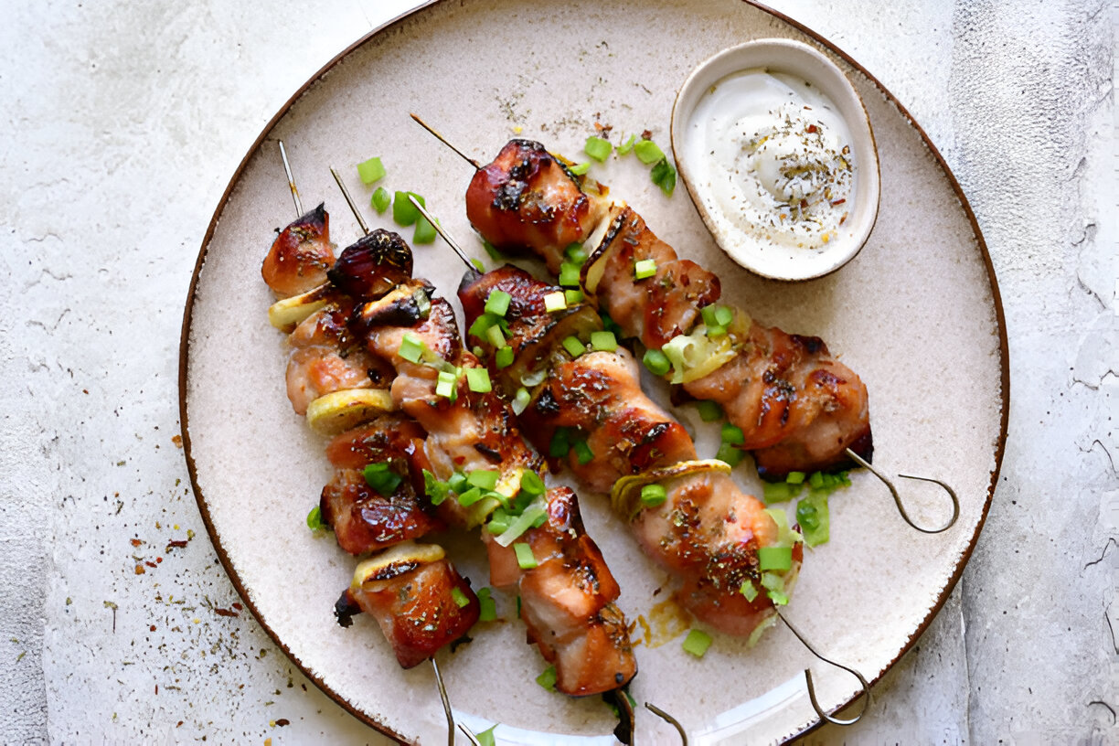 Grilled chicken skewers topped with green onions served with a yogurt sauce on a beige plate.