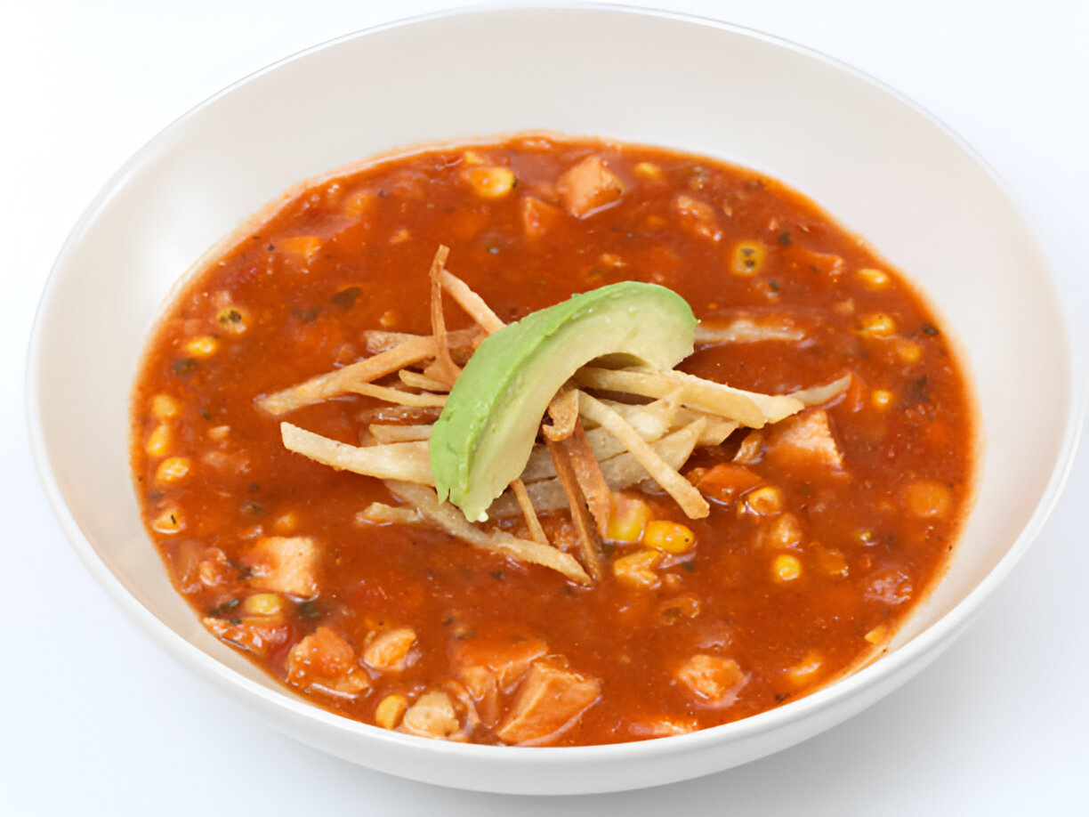 A bowl of tomato-based soup with corn, chicken, and tortilla strips, garnished with slices of avocado.