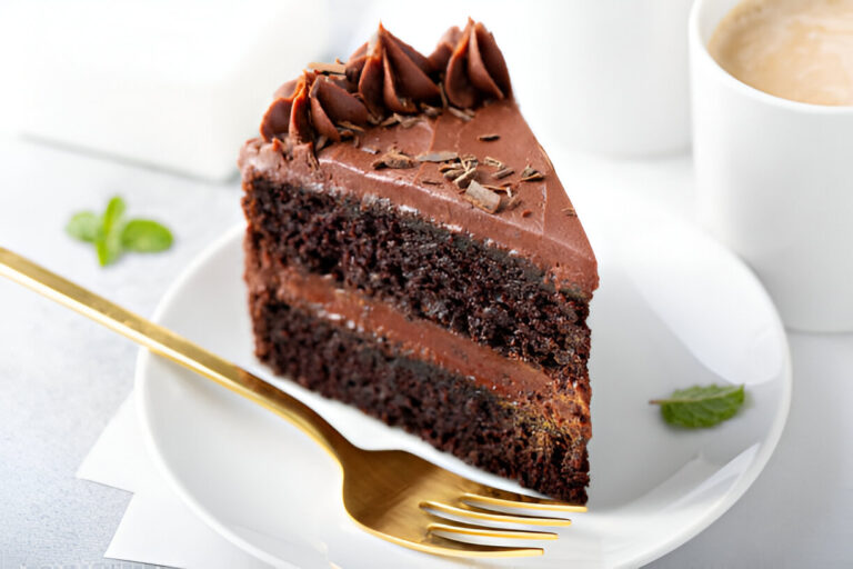 A slice of rich chocolate cake with chocolate frosting and a gold fork, served on a white plate with a mint leaf garnish.