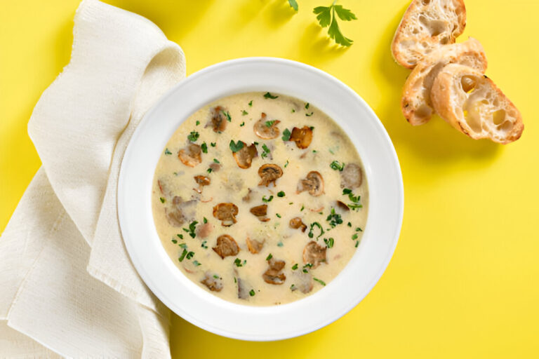 A bowl of creamy mushroom soup garnished with chopped parsley, alongside crispy bread slices on a bright yellow background.
