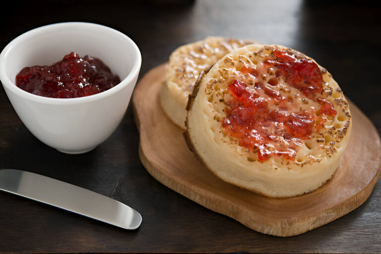 A delicious serving of toasted crumpets topped with strawberry jam on a wooden plate, accompanied by a small bowl of more jam and a butter knife.