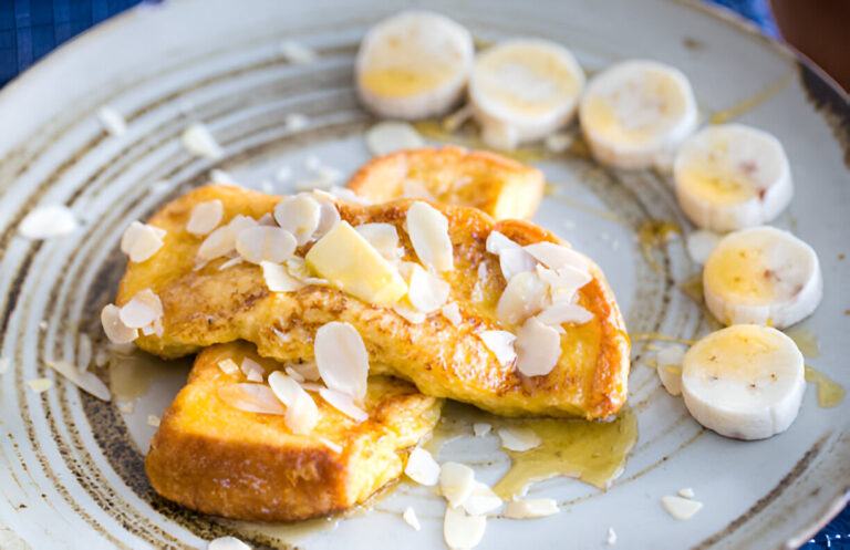A plate of French toast topped with sliced almonds and banana slices, drizzled with syrup.