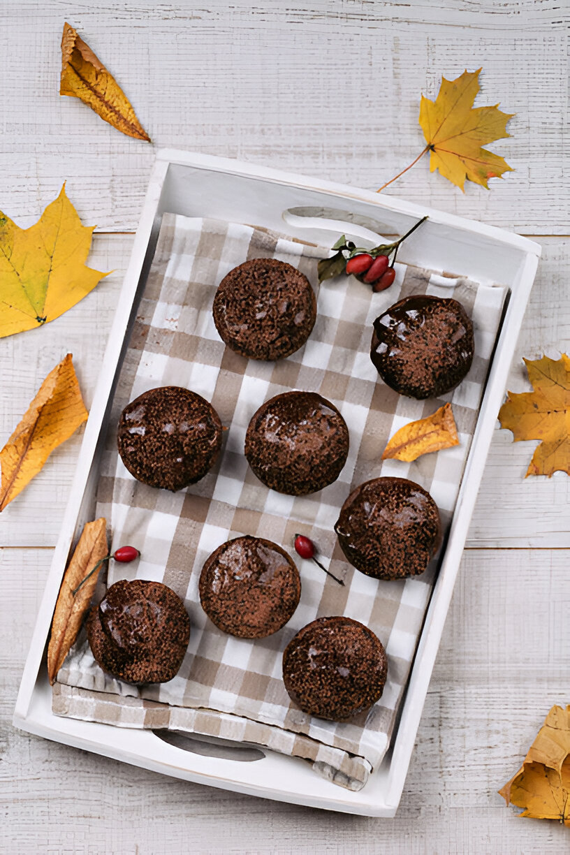 A white tray holding freshly baked chocolate muffins on a checkered cloth with autumn leaves scattered around.