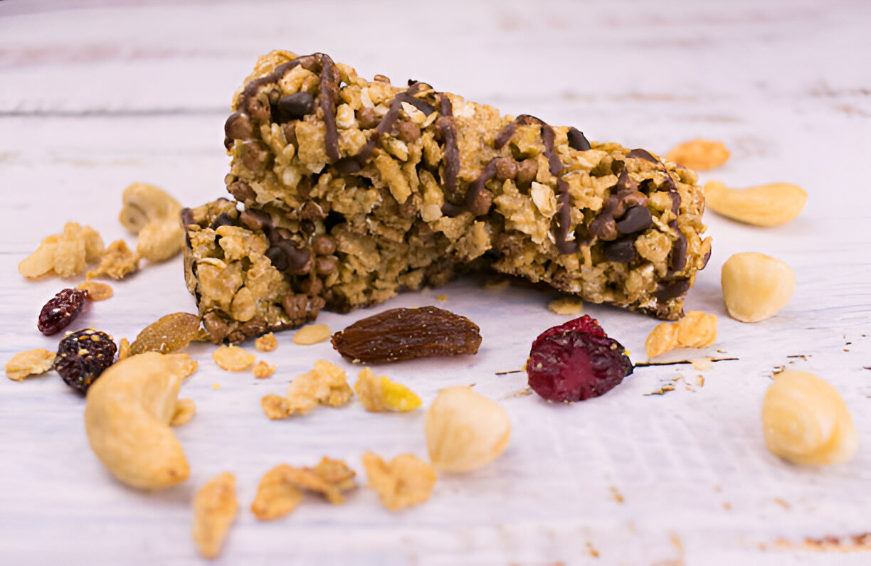 A close-up image of crunchy granola bars topped with chocolate drizzle, surrounded by mixed nuts and dried fruits.
