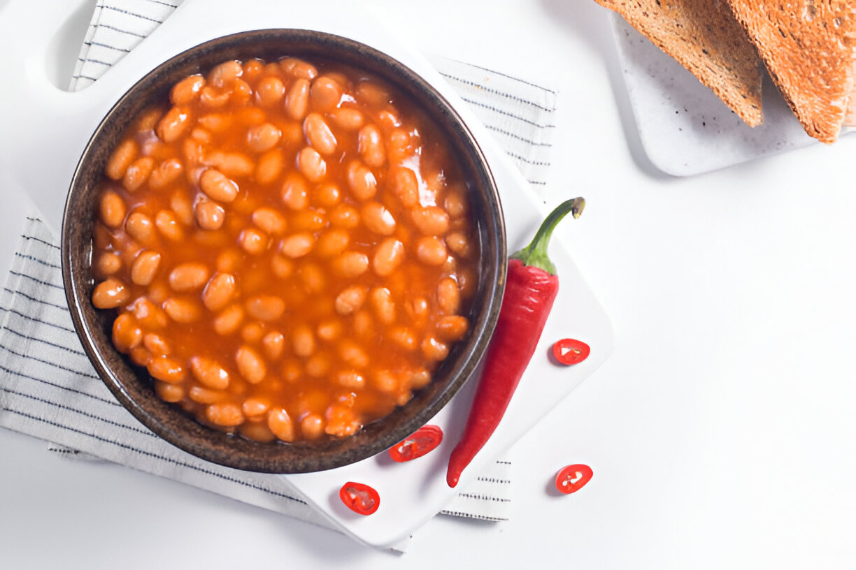 A bowl of baked beans in tomato sauce, surrounded by fresh red chili peppers and slices of toasted bread.