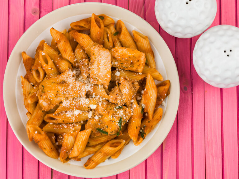 A bowl of pasta with a creamy orange sauce and grated cheese on top, next to white golf ball-shaped salt and pepper shakers on a pink wooden table.