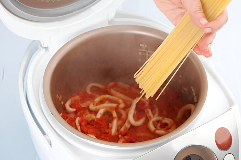 A hand holding uncooked spaghetti over a pot of cooking pasta in a rice cooker or multicooker.