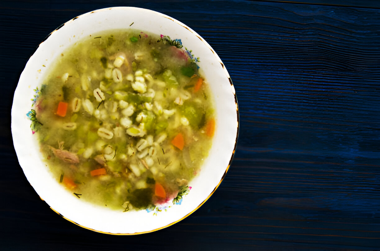 A bowl of soup with barley, vegetables, and herbs on a dark wooden table.