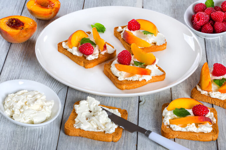 Slices of crisp bread topped with cream cheese, fresh peaches, raspberries, and mint leaves, on a wooden table.