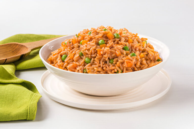 A bowl of colorful rice with peas and carrots on a white plate, surrounded by a green napkin and a wooden spoon.