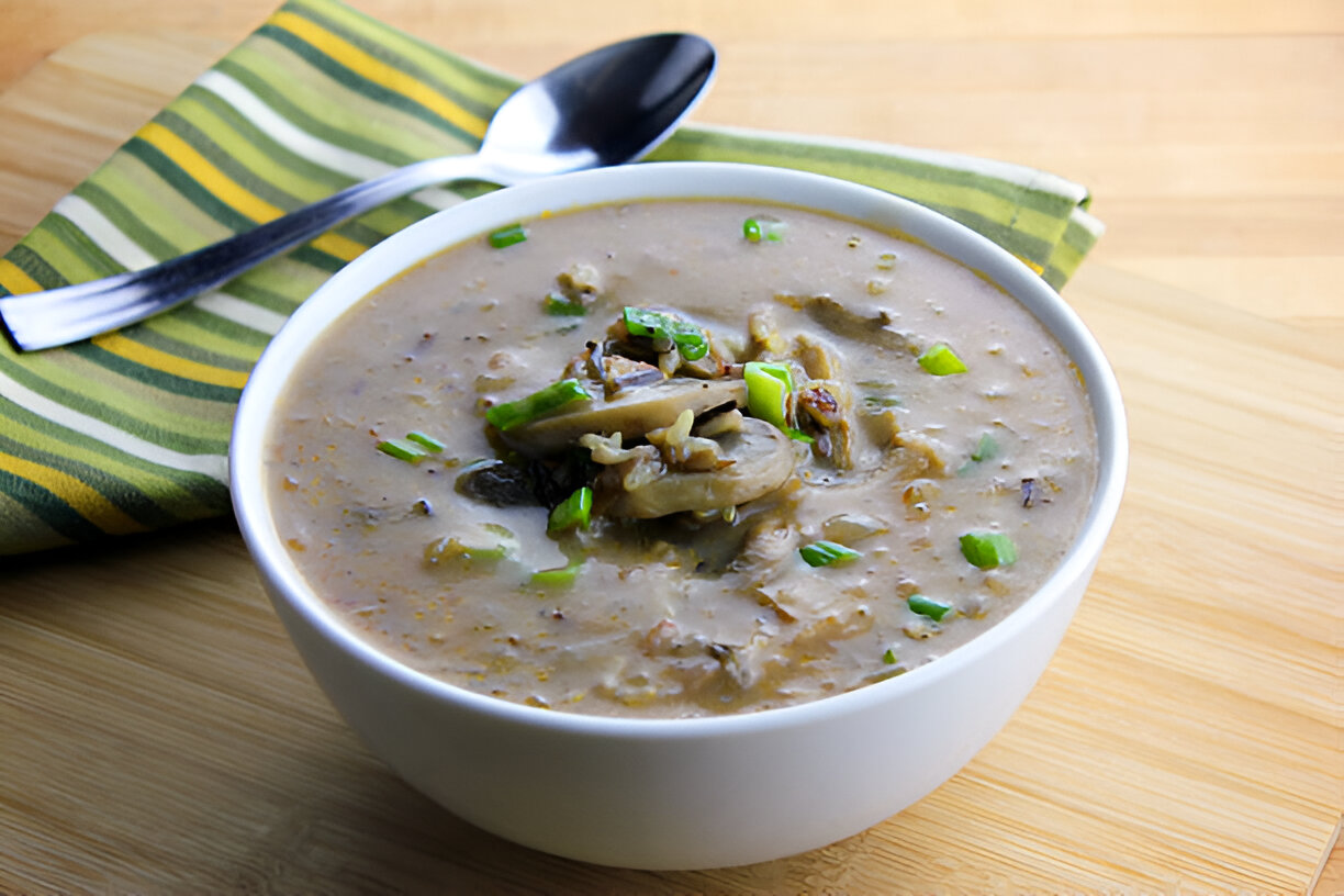 A bowl of creamy soup topped with sliced mushrooms and green onions, sitting on a wooden surface next to a striped napkin.