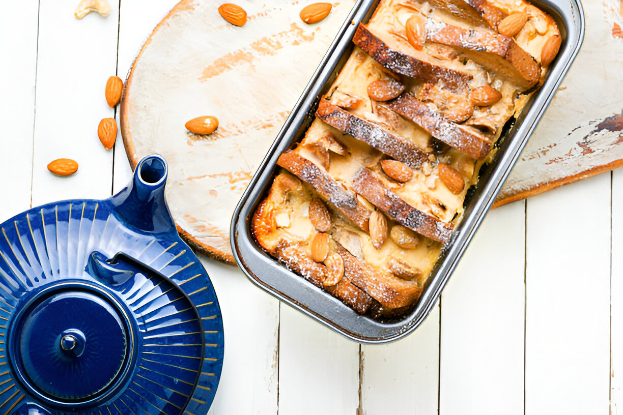 Delicious baked almond bread pudding in a metal pan, surrounded by scattered almonds and a blue teapot on a wooden surface.