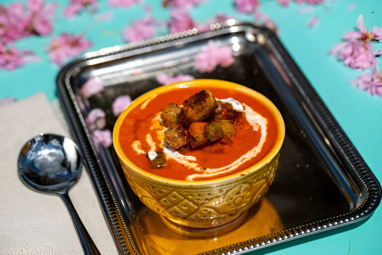 A vibrant bowl of red soup topped with crispy garnishes, surrounded by pink flower petals on a turquoise background.