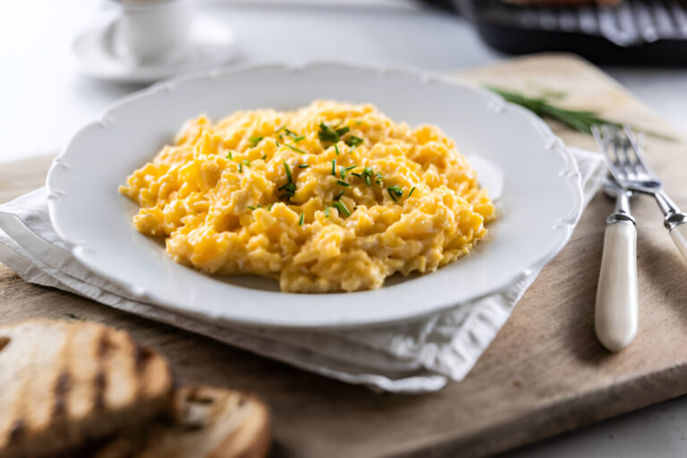 A plate of creamy scrambled eggs garnished with chives, served on a wooden board with toasted bread in the foreground.