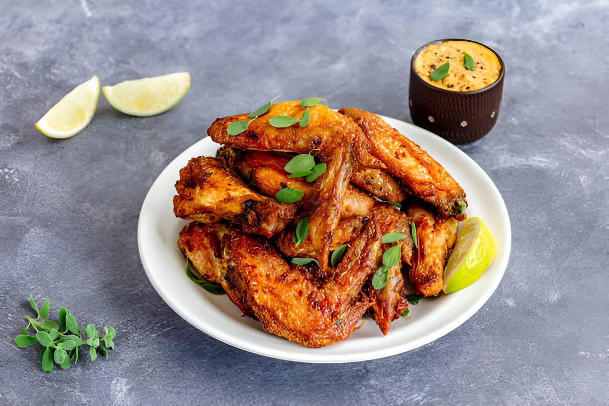 A plate of crispy chicken wings garnished with green herbs and a lemon wedge, accompanied by a small cup of dipping sauce.