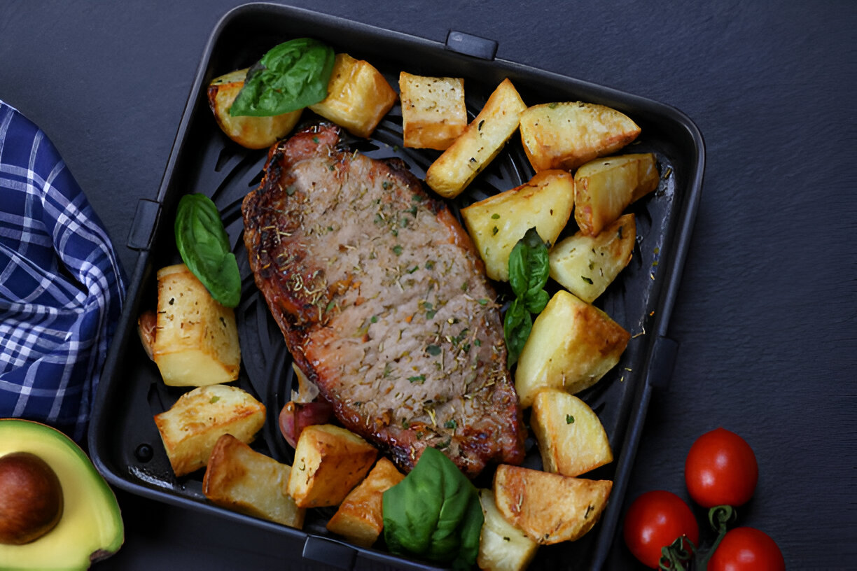 A delicious oven-roasted steak with crispy potatoes and basil leaves on a black tray, accompanied by cherry tomatoes and a knife and fork in a checked napkin.