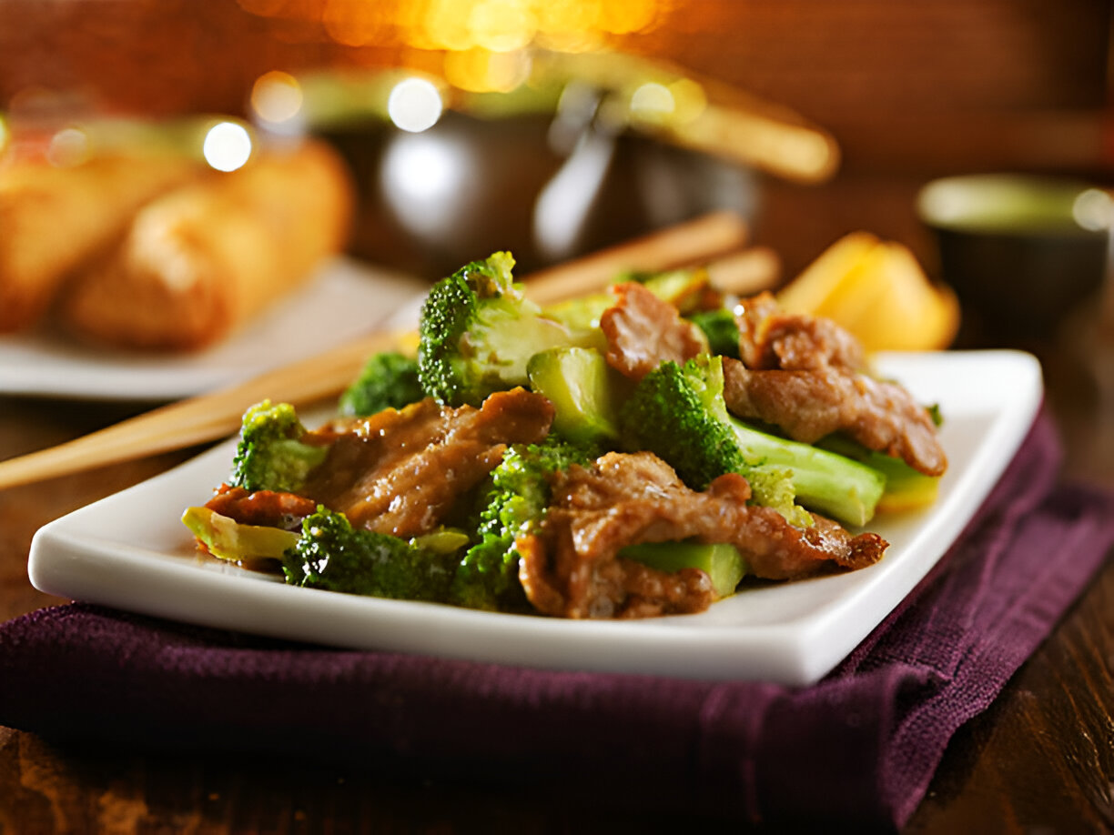 A plate of broccoli and beef served with chopsticks, with egg rolls in the background.